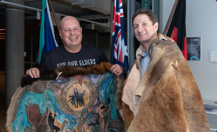 Alex Kerr and Ashley Firebrace holding a possum-skin cloak featuring their Innovate RAP artwork
